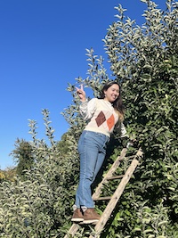 Emily wearing a white and orange argyle sweater while standing on a ladder in an apple tree.