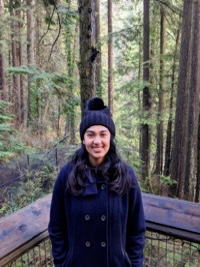 A headshot of Krithika in a black coat standing at a wooden railing with trees behind her.