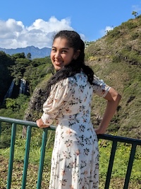 An image of Krithika wearing a white dress standing at a metal railing with a waterfall in the background.