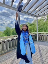 Sana wearing graduation regalia and smiling, arm holding her graduation cap high.