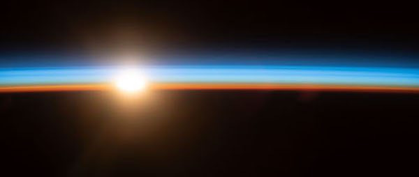 From space, the sun appears over Earth's horizon, illuminating the atmosphere as a blue layer above Earth. Above the atmosphere, space appears black.