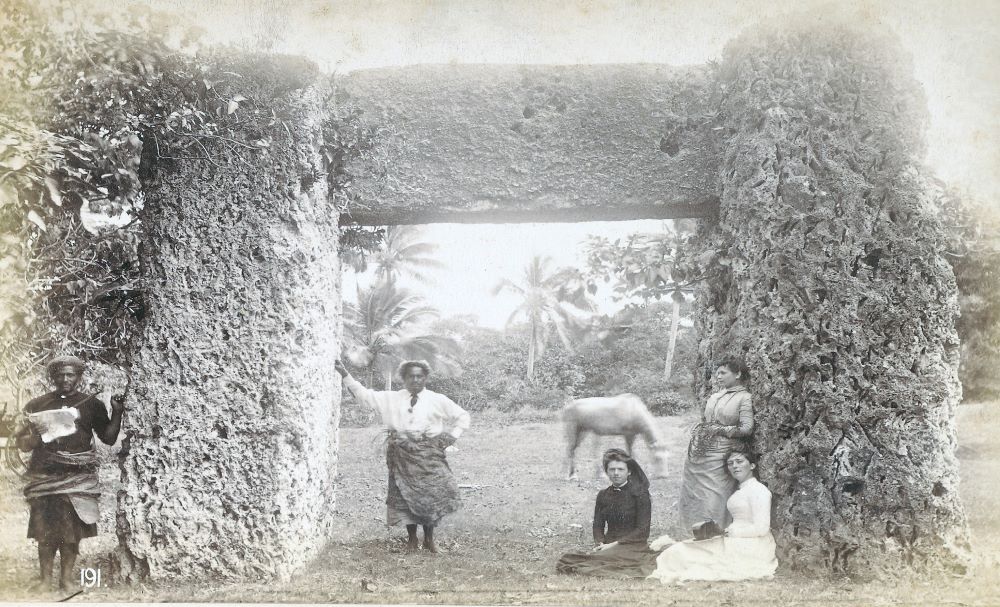 Photo of a large stone gateway consisting of two posts and a beam. Five people are pictured sitting and standing in the grass around and under the gateway.