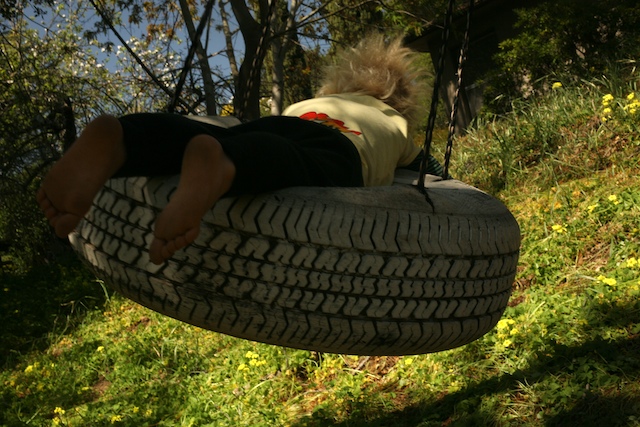 Photo of young child on tire swing with his feet facing the camera. The swing is near the high point of its trajectory and tilted away from the camera/viewer. 