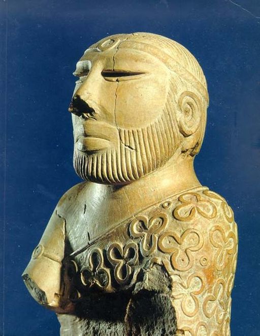 A steatite statue against a blue background. The statue depics a man with a beard and a headpiece wearing decorative dress across one shoulder.