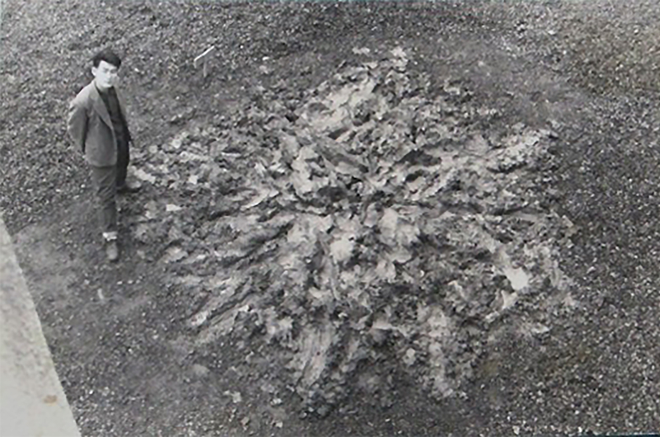 A pile of mud bearing an array of marks—indentations, cavities, and smears—sits at the center of a gravel field shot from a birds-eye view. A man, fully clothed in a casual suit, stands at left with hands behind him, head turned to directly face the camera following the convention of photographing painters next to their finished canvases. Shiraga Kazuo, Challenging Mud. (Doro ni idomu), 1955 (3rd execution). Postcard © Shiraga Fujiko and Hisao and the former members of the Gutai Art Association