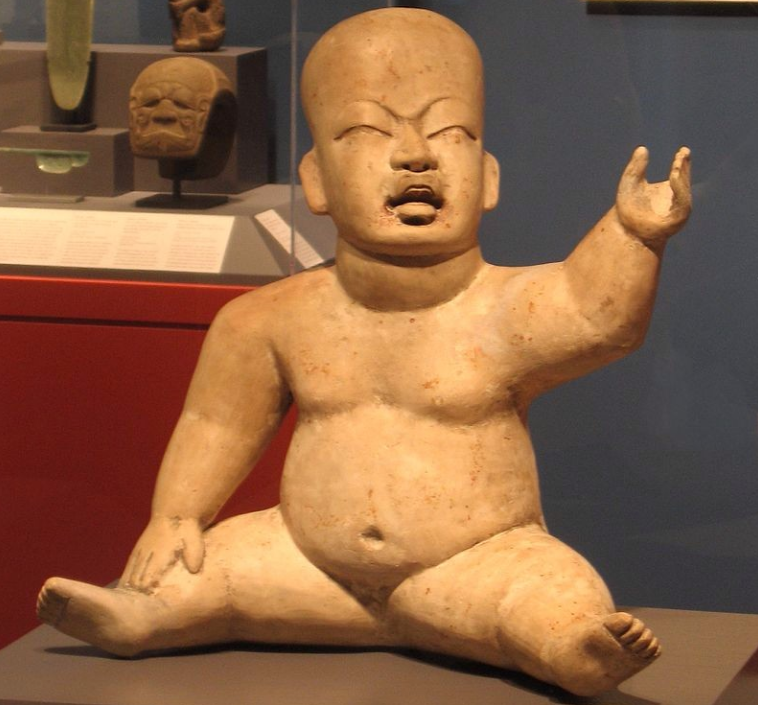 Photograph of a realistic ceramic baby figurine. The child is nude, with its eyes shut and furrowed brow, appearing to cry. It is seated and reaches one arm up. Its legs are splayed out.