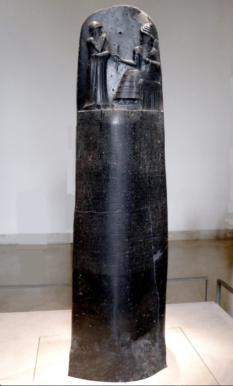 Law Code of Hammurabi inscribed on a black stone slab, rounded at the top and rectangular at the bottom. At the rounded top of the slab, taking up about a quarter of the space on the front, is a relief sculpture of two people, one sitting in a throne and wearing an elaborate gown, the other standing with their arms crossed. The lower portion of the slab has law codes written on it in cuneiform.