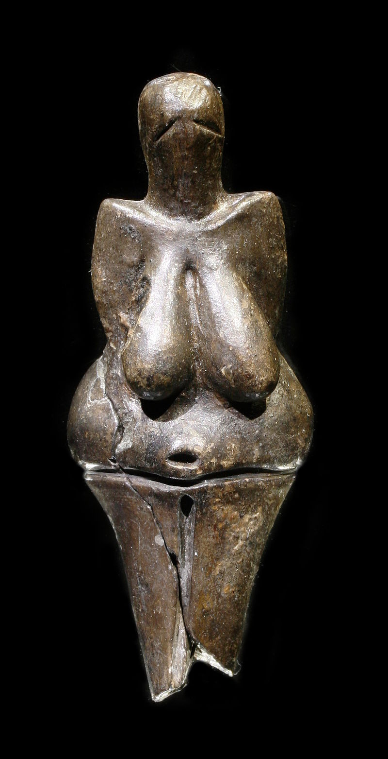 A statue made of polished dark stone representing a female figure with exaggerated breasts and hips. There are no defined facial features and distinct arms are not visible.