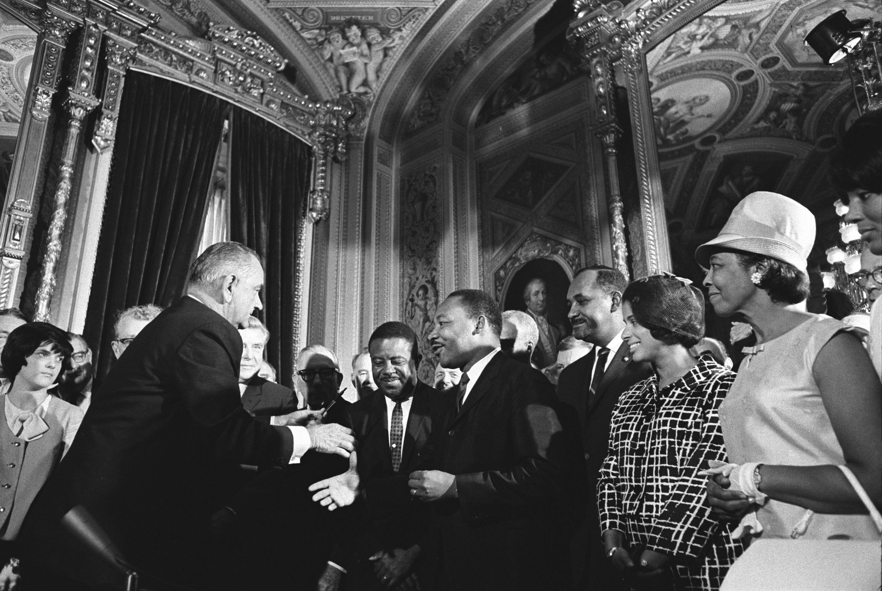 Black and white photograph of Lyndon Johnson extending a hand to Martin Luther King Jr. They are surrounded by a crowd of onlookers, both black and white.