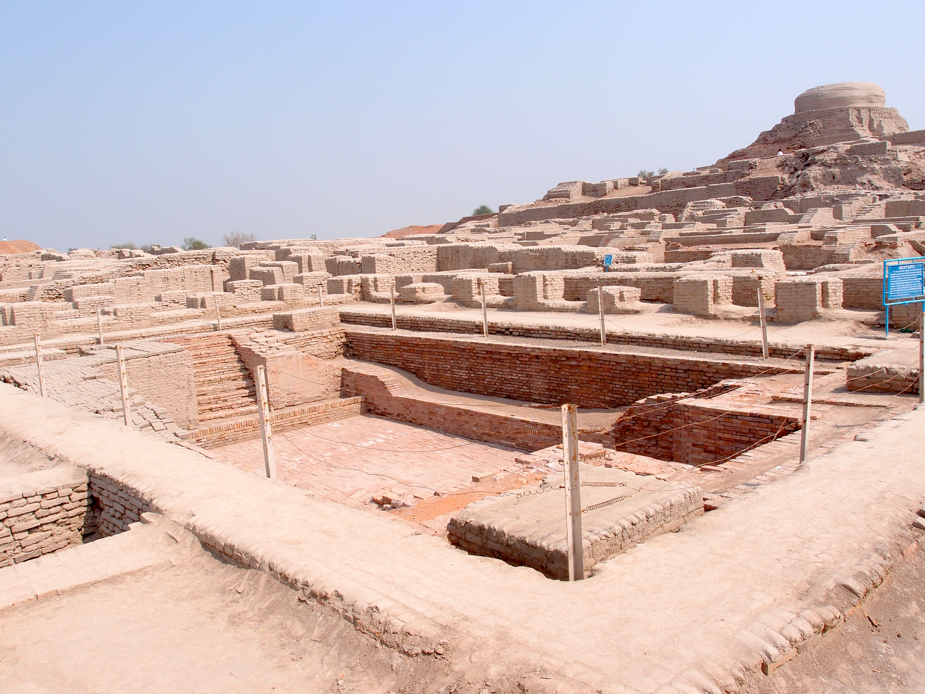The Great Bath at Mohenjo-daro: Amid the brick ruins of a 3rd-millennium BCE city, stairs descend on two sides into a large, rectangular brick-lined pit.  Wooden stakes and wire encircle the perimeter, preventing entry by modern-day tourists.